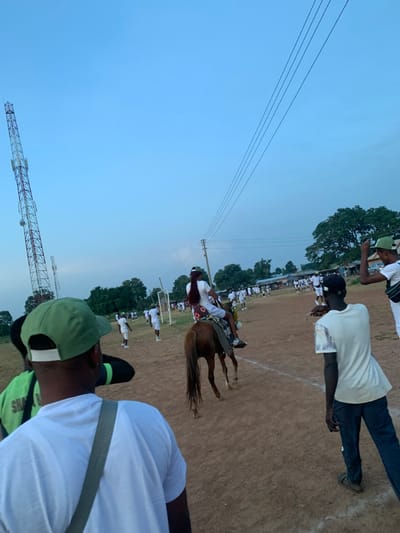 The image depicts an outdoor gathering on a reddish-brown dirt field in Kubi A, Nigeria, under a clear, light blue sky indicative of late afternoon or early evening. In the immediate foreground, facing away from the camera, is a male subject wearing a white short-sleeved t-shirt, a green baseball cap, and a grey shoulder strap. To his left, part of another individual in a green shirt is visible.
In the midground, a brown horse with a long tail is ridden by a person, whose back is largely to the camera. The rider wears a white top and has long, reddish-brown hair; the horse is adorned with a patterned saddle blanket. The horse and rider are moving from left to right across the frame. To the right of the horse, another male subject, also facing away, wears a light-colored t-shirt, dark trousers, and sandals. Further to the right, a fourth individual, wearing a white top and a green cap, is visible raising their right arm.
The background features numerous individuals, estimated between 50 and 70, mostly dressed in white or light-colored attire, spread across the field. Many appear to be standing or walking, some in loose formations. Two white soccer goalposts are visible in the distance, indicating the field's use. Behind the field, a line of green trees and faint outlines of structures are present. A tall, red and white striped lattice telecommunications tower dominates the far left of the background, while wooden utility poles carrying multiple black power lines extend across the upper right portion of the scene.