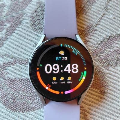 The image shows a close-up view of a smartwatch with a light purple band. The watch face displays the time "09:48", a Bluetooth icon with "BT 23", a battery indicator, weather icons, a percentage indicating "55%", and a gauge with numbers and colored segments. There are also text labels "10AM 11AM 12PM", "112", a sun icon, and the word "Низкий" which is Russian for "Low". The watch is placed on a textured fabric surface, possibly a tablecloth or a blanket, with a pattern of interwoven threads in shades of white, beige, and pink. The image is taken from a slightly elevated angle looking down. The scene is indoors and appears to be a still life composition focused on the smartwatch. There is no indication or context of the location being Skuķīši, Latvia.