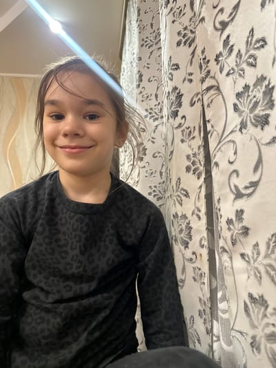The image is oriented sideways, appearing rotated 90 degrees clockwise. It features a young girl, likely pre-teen, looking upwards with a gentle, cheerful smile directed at the camera. Her light brown hair is loosely pulled back, with a few strands framing her face. She is wearing a dark, possibly black or very dark grey, long-sleeved top featuring a subtle, tone-on-tone leopard print pattern.
Above and slightly behind her, a light-colored fabric with a repeating grey and white floral pattern is prominent, suggesting curtains or bedding. Detailed crops reveal that this fabric might include a mesh layer beneath the patterned material. A strong, bright light source is visible to the girl's left, creating a significant lens flare and illuminating her face. The overall setting appears to be indoors, likely a home environment. No visible text is present in the image. The girl's expression conveys a sense of happiness and ease as she poses for the photograph.