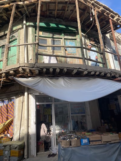 This image depicts a street scene in Kabul, Afghanistan, likely a marketplace given the shops and goods on display. The architecture suggests older construction with wooden beams, balconies, and weathered paint, particularly on the upper levels of the buildings.

In the foreground, a market stall is set up with a variety of goods spread out on tables, covered by a grey tarp. Prominently displayed are numerous padlocks of various sizes, arranged in cardboard boxes and baskets. Other items visible include what appear to be tools or hardware, and possibly some packaged goods. To the left, a store window shows shelves stocked with boxes, and two people are visible inside. One person is seated, seemingly attending to business, while another is partially visible in the background.

Further left, a colorful striped fabric or banner hangs, and stacks of wrapped packages, likely merchandise, are piled up. Above the market stall, the facade of an older building is visible, with green painted doors and windows. A balcony area is present, supported by wooden posts. On this balcony, a man wearing a light blue garment and a cap is leaning over, engaged in a conversation on a mobile phone.

The overall impression is of a busy urban environment with a mix of commerce and residential or commercial structures, characteristic of many areas in Kabul. The lighting suggests it is daytime, with shadows indicating the sun's position.