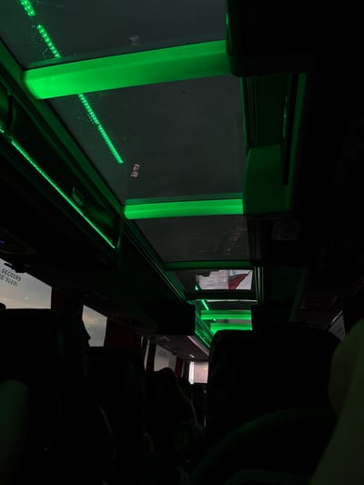 The image depicts the interior of a bus, likely on a journey, with a prominent feature being the ambient green lighting that illuminates the ceiling and reflects off the windows. The perspective is from a passenger's seat, looking upwards and forward.
The ceiling of the bus is equipped with multiple strips of green LED lights, casting a vibrant glow. Above the seats, there are overhead compartments, and some of these are also lit with the same green hue. Through the transparent sections of the ceiling, what appears to be a flag can be seen reflecting, or possibly being displayed on a screen.
The seats themselves are largely in shadow, suggesting the main light source is the artificial green lighting. The windows are visible, some showing a cloudy sky, hinting at the time of day or weather conditions outside. Text is visible on a window in the lower left, reading "SECOURS" and some numbers, likely related to emergency exits or instructions.
There are no discernible individuals clearly visible in the image due to the darkness and focus on the lighting and interior architecture. The overall impression is one of a modern bus interior, possibly designed for night travel or to create a specific atmosphere. The location in the city of Siret, Romania, is not visually indicated within the image itself.