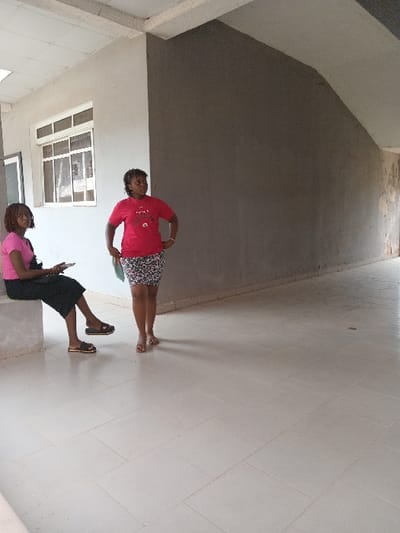 This image depicts two adult females within an indoor or semi-enclosed common area of a building in Ogboko, Nigeria. The floor is laid with light-colored square ceramic tiles. Walls are painted in two distinct tones: a lighter grey/off-white on the left, which includes white-framed windows, and a darker grey on the right. Overhead, a suspended ceiling with white tiles and structural beams is visible, alongside angled architectural features.
On the left, one female is seated on a light-colored concrete bench. She is dressed in a pink short-sleeved top, a dark skirt, and light brown flat sandals. She holds a dark electronic device in her hands and directs her gaze downwards. A dark bag rests on the bench beside her.
Standing slightly forward and to the right is a second female. She wears a red short-sleeved t-shirt with a graphic design, a patterned short skirt, and light brown flat sandals. Her hands are positioned on her hips, and she holds a light-colored object in her left hand, looking towards the left side of the frame. There is no direct interaction observed between the two individuals.