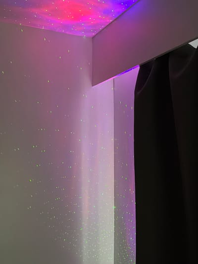 The image depicts a corner of a room illuminated by a projector displaying a nebula and star effect on the ceiling and walls. The ceiling features a colorful nebula with shades of pink and purple, interspersed with small green dots representing stars. The walls are predominantly white but tinted with a soft purple hue from the projector's light, and they are also dotted with the same green star-like lights. A dark, solid curtain occupies the right side of the frame, obscuring a portion of the room. The perspective is from a low angle, looking upwards towards the ceiling. No people or specific landmarks indicating a location in Turnhout, Belgium are visible.