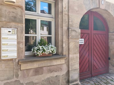 The image shows a section of a stone building in Erlangen, Germany. A white mailbox with multiple compartments is mounted on the left side of the wall. To the right of the mailbox is a window with white frames, showing reflections of the sky and surrounding buildings. A planter filled with white flowers sits on the windowsill. To the right of the window is a large, arched red wooden door. A white sign with German text, likely a warning or instruction, is attached to the wall to the left of the door. The cobblestone ground is visible in the foreground.