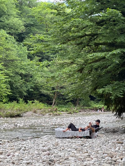 Two people are relaxing by a rocky riverbank, with a forest-covered mountain in the background. The people are on a low-lying cot or bed. One person is lying down, wearing black leggings and a black top, with their head propped on a pillow. The other person is sitting, wearing a black baseball cap and black t-shirt, and appears to be looking at a phone. The river is shallow and full of smooth, grey stones. The surrounding vegetation is lush and green, with trees creating a canopy over parts of the scene. In the distance, other people are visible further up the riverbank.