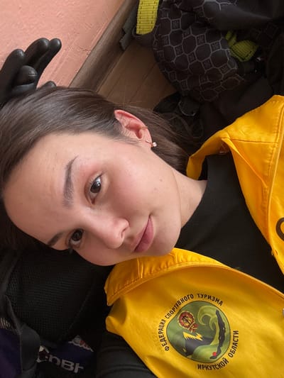 A female individual is depicted in a close-up, angled shot, appearing to recline indoors. She has dark hair, a fair complexion, and brown eyes, looking directly at the camera with a slight expression. Her right ear displays a small, light-colored stud earring. A black glove covers her partially visible left hand, positioned near her head. She wears a black long-sleeved garment underneath a bright yellow zippered vest. The vest features a circular emblem on the left chest, displaying the Russian text "ФЕДЕРАЦИЯ СПОРТИВНОГО ТУРИЗМА ИРКУТСКОЙ ОБЛАСТИ" (Federation of Sports Tourism Irkutsk Region), encircling a multi-colored logo depicting a mountainous landscape. Partially visible behind her are a black object with a mesh texture, and another dark object with a geometric hexagonal pattern. A light orange wall and a segment of a wooden floor or trim are visible in the background. The scene is consistent with an indoor setting within the Irkutsk Region, Russia, given the vest's insignia; however, the precise location within the city of Tayshet, Russia, is not discernible from the image.