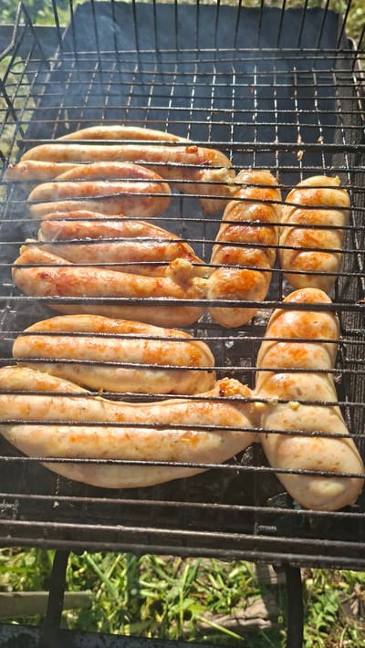 The image shows several sausages grilling on a metal grill. Smoke is rising from the grill. The sausages appear to be partially cooked with some browning visible. Some grass is visible around the grill. The location is near Сваткаўскі сельскі Савет, Belarus.
