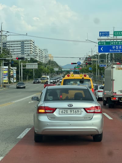 The image shows a street scene in Nonsan-si, South Korea, likely during the daytime given the lighting. A silver Kia sedan is in the foreground, facing away from the camera. The car's license plate reads "62도 9169". The car is in a traffic lane with red pavement markings. Several other vehicles, including a yellow school bus, a white truck, and other cars, are visible on the road, indicating moderate traffic. Traffic signals and road signs are present, including a sign pointing to "Nonsan Tax Office" and "Buchang-ro". Apartment buildings and trees line the street, suggesting an urban or suburban environment. The sky is overcast with some clouds.
