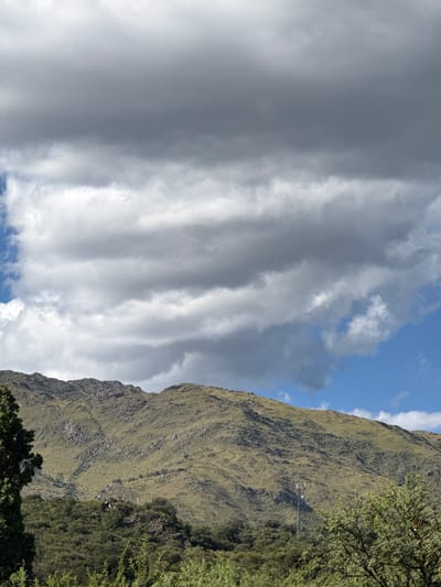 A landscape featuring a lush, grassy mountain partially obscured by leafy trees. Beside the mountain, a sky filled with long, undulating layers of gray clouds dominates the frame, with a glimpse of blue sky visible between the clouds and the mountain. A small utility tower can be seen on the mountain side.