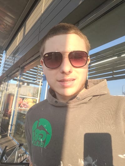 A young male subject, appearing to be in his late teens or early twenties, is depicted in a selfie-style photograph taken in Kuurne, Belgium. He has short, light brown hair and is wearing aviator-style sunglasses with dark, gradient lenses and a gold-colored frame featuring a crocodile logo on the left temple. He is dressed in a dark gray or olive-green hooded sweatshirt with a partially obscured green circular logo on the left chest, which appears to read "BUILD". The sweatshirt exhibits multiple light-colored paint or stain marks. Strong sunlight casts a distinct shadow diagonally across his face and torso. The subject is positioned outdoors in front of a modern building entrance, likely a supermarket or grocery store, characterized by large glass panels. To the subject's left, a stack of metal shopping carts is visible. Through the glass, an interior display of fruits, including grapes, apples, and oranges, can be seen. A metallic canopy extends overhead above the entrance.