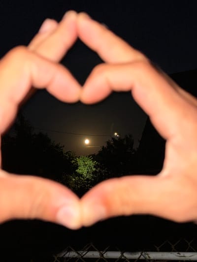 The image shows a close-up of two hands forming a heart shape, with a night sky and trees visible through the center. A bright circular object, likely the moon, is visible in the sky. There are also two small points of light in the sky, possibly streetlights or distant stars. The foreground shows a chain-link fence. The image appears to be taken at night in a semi-urban or rural setting, as indicated by the presence of trees and a fence, and the bright celestial object. The location in Радашковіцкі сельскі Савет, Belarus is not visually evident from the image itself.