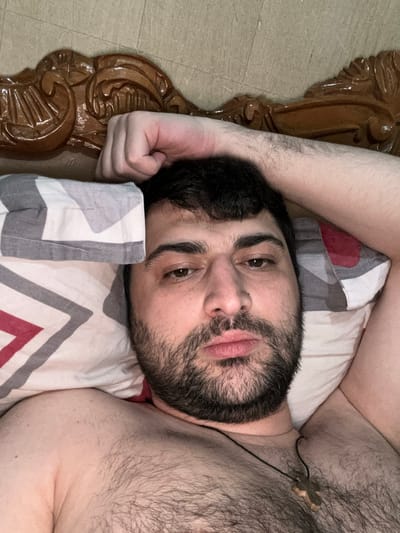 The media file shows a close-up, overhead shot of a person lying on a bed. The person is a male with dark hair and a beard, wearing a necklace with a cross pendant. He is lying on his back with his head resting on a pillow that has a geometric pattern of red, white, and grey stripes. His left arm is bent at the elbow, resting on the pillow, and his right arm is not fully visible. The person's eyes are open and looking upwards, and his expression appears neutral or relaxed.

To the left of the frame, a portion of a wooden bed frame with intricate carvings is visible. The bed frame is dark brown and appears to be made of polished wood. The flooring around the bed is a light-colored tile.

The lighting suggests it could be daytime, perhaps indoors in a dimly lit room, or early morning or late evening. There are no overt clues about the weather. The presence of the cross pendant might suggest a religious affiliation or personal significance. The overall scene depicts a moment of rest or waking up in a bedroom. The image does not contain any visible text.