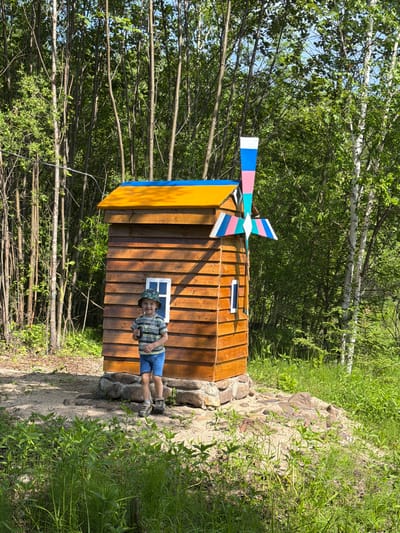 A young boy with a bucket hat, striped t-shirt, and blue shorts stands in front of a small, wooden windmill structure. The windmill has a yellow roof with a blue trim and a colorful pinwheel. The structure is surrounded by rocks and sandy ground, with lush green vegetation and trees in the background. The scene appears to be in a park or outdoor recreational area in Kul'dur, Russia, evidenced by the birch trees and the overall natural setting. The lighting suggests it is daytime, with sunlight filtering through the trees.
