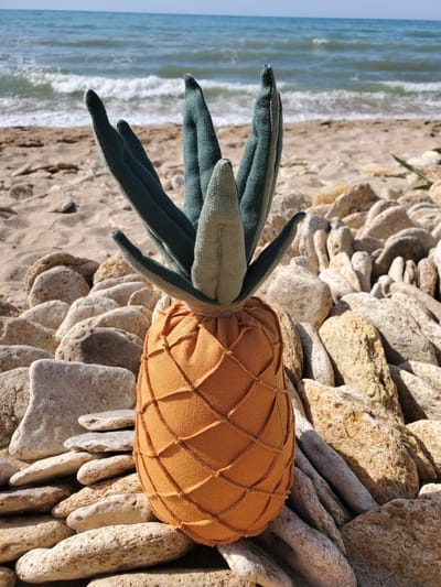 A plush, fabric pineapple toy sits on a rocky beach, with the Black Sea in the background. The toy is positioned in the center of the frame, its orange, patterned body resting on a bed of smooth, light-colored stones. Green, leaf-like fabric pieces extend upwards from the top of the pineapple, mimicking its natural crown. The beach is composed of sand and numerous rounded stones of varying sizes and colors, ranging from white to tan and grey. The sea, a deep blue, stretches across the horizon, with gentle waves creating white foam as they approach the shore. The sky above is a clear, pale blue. The scene is set during daylight, with sunlight casting shadows on the rocks and the toy.