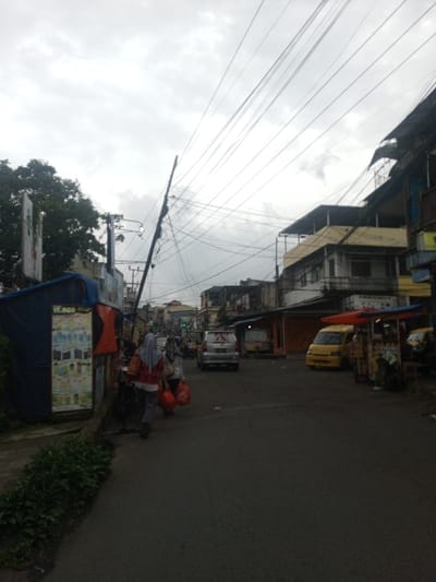 The image depicts a street scene in Sindanglaya, Indonesia, under an overcast sky. The view is along a paved, dark asphalt road, with buildings and commercial structures lining both sides.
On the immediate left, a blue tarp-covered stall, accented with orange fabric, displays merchandise on a visible poster advertising "Rp 1.000". Above this, dense green tree foliage is visible, alongside a utility pole laden with numerous intertwined electrical and communication cables extending across the frame. Two individuals are walking away from the viewer on the left side of the road. The leading person wears a light-colored hijab, a red top over a white shirt, grey pants, and carries multiple red plastic bags in their left hand. A second person, also in a light-colored hijab, walks behind them. A strip of green ground cover or bushes lines the immediate left edge of the road.
On the right side of the road, multiple multi-story buildings, some appearing residential-commercial with balconies and various roofing types, are visible. A yellow van, possibly a public transport vehicle, is parked or stopped near a red and orange canopied street vendor stall. Further down the street, a silver minivan or SUV is visible, heading away from the camera. Additional vehicles and indistinct figures are present further in the distance. Overlapping power lines dominate the upper half of the frame against the bright, cloud-filled sky.