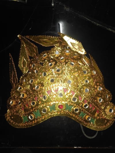 The image presents a golden, ornate headpiece, reportedly located in Melreux, Belgium. The object, appearing to be a crown or ceremonial mask, is predominantly gold-colored and constructed from a reflective, possibly metallic or foil-like material. Its curved surface is heavily embellished with numerous small, circular, mirror-like elements. Stylized, pointed, leaf-like protrusions extend from the upper edges of the headpiece. Along the lower, wavy border, a green band with embroidered or painted patterns is visible, accented with small golden beads and a central, prominent red circular gem or bead. Additionally, scattered red or pinkish triangular motifs adorn the main golden body. A thin, light-colored elastic string is partially visible extending from the bottom right side. The headpiece rests on a dark, reflective surface, likely black, which shows minor surface scratches, dust, and fine linear strands, consistent with faint cobwebs or surface imperfections. No individuals or other subjects are present in the scene.