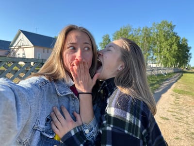 Two young women are depicted in a close-up outdoor selfie, presumably taken in Ust'-Silayka, Russia, under a clear, bright blue sky. The woman on the left, with blonde hair, wears a light blue denim jacket over a dark pink garment. Her right hand covers her mouth, and her eyes are wide with a startled or surprised expression, looking towards the right side of the frame. A small black graphic is visible on the upper left pocket area of her denim jacket, and a yellow object is partially visible in the jacket's lower left pocket. The woman on the right, with brownish-blonde hair, is wearing a dark blue and green plaid flannel shirt. She is leaning towards the first woman, mouth wide open as if yelling or laughing, with her eyes closed or squinted. Her right hand is placed on the first woman's left shoulder area. She has visible freckles and a black hair tie on her left wrist.

The background on the left includes a light-colored wooden lattice fence. Behind the fence, a light pink building with a dark blue pitched roof and at least one white-framed window is visible, with a wooden support structure leaning against its gable. A second similar building is partially obscured behind it. On the right side of the frame, a dirt path or road recedes into the distance, flanked by a rustic wooden fence and a dense line of green-leafed deciduous trees, consistent with birches. The scene is illuminated by direct sunlight, casting shadows.