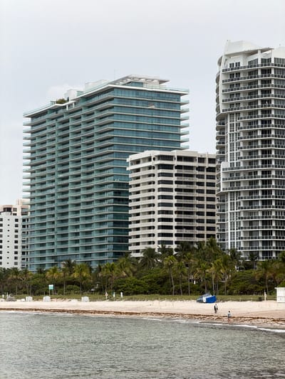 The image depicts a beach scene in Bal Harbour Village, United States of America, with three high-rise buildings in the background. The building on the left is a contemporary, multi-story structure with a facade primarily composed of horizontal bands of glass and white framing. To its right is a shorter, white building with a grid-like pattern of windows and balconies. The tallest building on the right is a modern residential tower with a curved, white exterior and numerous balconies with dark railings.
In the foreground, a sandy beach stretches across the lower portion of the image. The sand is light-colored and has some patches of seaweed. Several lounge chairs, neatly stacked, are visible on the beach, along with some small white tables. A blue tarp covers a boat or structure near the tree line. Two individuals are walking on the beach near the water's edge. One appears to be a man in a dark jacket and light pants, and the other is a woman in shorts and a light-colored top. Gentle waves lap at the shore. A line of palm trees and lush green foliage separates the beach from the buildings. The sky is overcast and pale, suggesting a cloudy day.