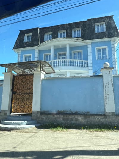 An exterior daytime shot in Pesochny, Russia, captures a two-story light blue house with a dark gray mansard roof and white architectural trim. The house features numerous rectangular windows with white frames and grids on both main floors and within the dormers of the roof. A prominent curved balcony with white balustrades and supporting columns is centered on the second floor. In the foreground, a light blue solid wall fence with white decorative pillars runs parallel to the street. An ornate, golden-colored wrought-iron gate with intricate scrollwork is integrated into the fence on the left, beneath a curved, translucent canopy. The house number "97" is visible on a white plaque attached to the left gate pillar. The ground level includes a concrete sidewalk or driveway in the immediate foreground, with a strip of grass containing small yellow dandelions bordering the fence. The sky overhead is clear and bright blue, traversed by multiple dark power lines. No individuals or discernible actions are present within the scene.