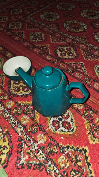A teal-colored, faceted teapot with a matching lid and handle sits on a red rug with an intricate geometric pattern. Next to the teapot, on the left side of the image, is a small, white bowl with a teal rim. The rug's pattern features repeating diamond shapes and floral motifs in shades of red, yellow, black, and white. The scene is lit from above, casting shadows on the rug. This image was taken in Gurlan, Uzbekistan.