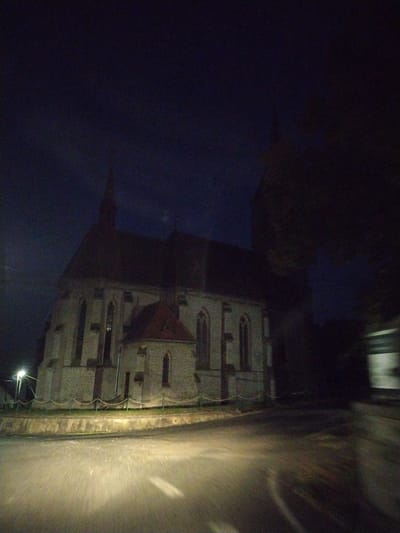 The image depicts a church, likely Saint Mary Magdalene Church, in the city of Mešno, Czechia, at night. The sky is dark blue and the church is illuminated by ambient light, possibly from streetlights and its own interior lighting, casting a pale glow on its stone facade. The architecture features Gothic-style arched windows and a pitched roof with a spire, parts of which are silhouetted against the night sky. A road is visible in the foreground, blurred due to motion, suggesting the photo was taken from a moving vehicle. A streetlight is visible to the left of the church, and a low fence with rope detailing separates the church grounds from the roadside. The overall scene is dark and somewhat indistinct due to low light conditions and motion blur, creating a somewhat atmospheric and moody impression of the building at night.