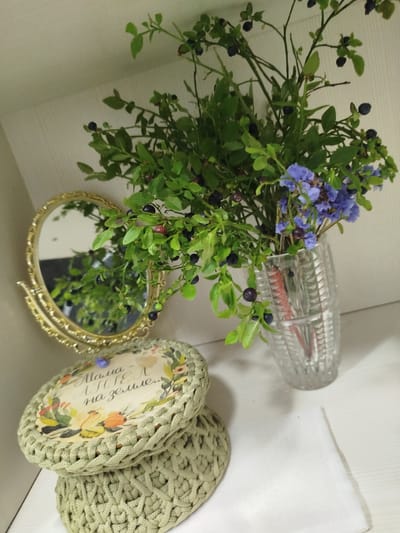 A still life composition features a bouquet of blueberry branches with small blue flowers and dark berries in a clear, faceted glass vase. To the left of the vase sits a gold-framed oval mirror on a stand, reflecting greenery. In the foreground, two crocheted, pale green baskets are stacked, the top one decorated with a round inset featuring a floral illustration and Cyrillic text that reads "Мама - АНГЕЛ на земле..." (Mom - an ANGEL on earth...). A white cloth is placed to the right of the baskets. The items are arranged on a white shelf, likely in a home setting. The overall scene is well-lit, with soft shadows. The location is described as Азарыцкі сельскі Савет, Belarus.