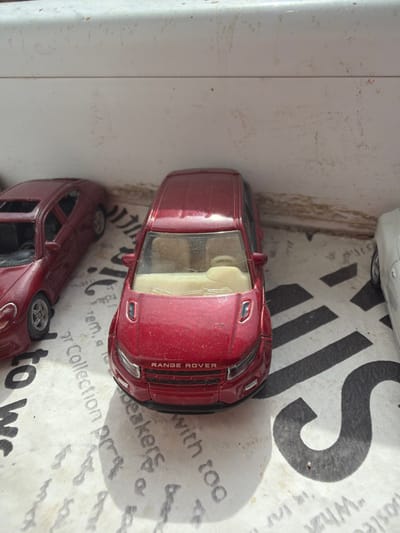 The scene displays three dusty metallic toy cars, set within the city of Kermen, Bulgaria. In the foreground, a prominent red metallic Range Rover Evoque model faces forward and slightly right, exhibiting significant dust accumulation on its exterior and a scuffed windshield. Its light-colored interior is visible. To its left, a partial view of a second red metallic toy sports car, resembling a coupe with a sunroof, is present, also showing dust and a silver multi-spoke wheel. To the far right, only the front-right portion of a third, silver or light-grey metallic toy car is visible. The vehicles rest on a flat, light-colored surface featuring black printed text and graphic patterns, including discernible fragments like "r Collection pr" and "speakers with too." The surface and an off-white vertical background behind the cars are notably grimy and covered with dust and debris. Bright overhead lighting casts clear shadows from the objects onto the patterned surface.