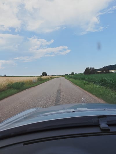 The image shows a view from inside a car looking out at a two-lane asphalt road. The car's hood and windshield are visible in the foreground, with a Mercedes-Benz emblem on the hood. The road extends into the distance, flanked by fields of grain and corn on either side. In the background, there are trees and a few buildings, suggesting a rural or semi-rural setting. The sky is partly cloudy, with blue sky visible between white clouds. The overall impression is that of a scenic drive through the countryside. It is not possible to determine the specific location of Obermodern-Zutzendorf, France, from this image.
