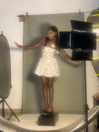 A young Black female is captured in a studio setting, posing dynamically. She is wearing a white dress with black polka dots and a delicate necklace. Her left arm is extended, and her right hand rests near her waist. Her left leg is bent at the knee, while her right leg is extended straight. The background is a seamless gray backdrop, indicating a professional photoshoot environment.
Lighting equipment is visible, suggesting an indoor studio shoot. The lighting appears to be artificial, with a prominent setup of panels with yellow accents in the upper left corner of the frame. The overall lighting is bright and evenly distributed, illuminating the subject and creating soft shadows.
A dark rectangular block is positioned beneath the subject's right foot, serving as a prop or platform to elevate her. To the right of the subject, a large, curved, reflective object, possibly a reflector or a piece of studio equipment, is partially visible. Another circular object, likely a softbox or umbrella, is present in the lower part of the frame.
The image is oriented vertically, with the subject appearing to be in motion or a contorted pose. The scene suggests a creative and artistic photoshoot, possibly for fashion or editorial purposes. The woman's expression is focused and engaged, hinting at a professional and confident demeanor. The overall ambiance of the image is clean and modern, characteristic of a controlled studio environment. The location context of South Ropp, Nigeria, provides a geographical setting for this photographic activity.
