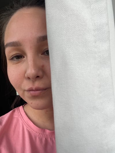 A woman is visible from the nose down, partially obscured by white fabric above. She has brown hair and eyes. She is wearing a pink shirt. The location cannot be determined from the image.