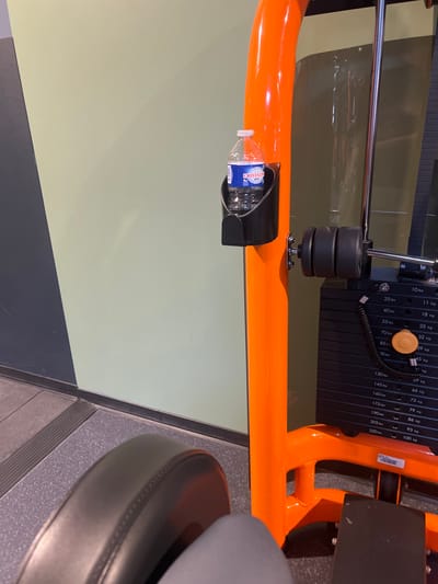 The image depicts an interior scene within a fitness center in Annœullin, France. A transparent plastic bottle of Cristaline brand water, featuring a blue label and pink cap, rests within a black holder attached to the bright orange, tubular frame of a gym machine. To the immediate right of the orange frame, a black weight stack is partially visible, displaying various weight increments in both pounds (e.g., 10 lbs, 25 lbs, up to 220 lbs) and kilograms (e.g., 5 kg, 11 kg, up to 100 kg). An orange selector pin is inserted into one of the weight plates, indicating a chosen resistance level. In the foreground, the dark grey padded section of another piece of exercise equipment is visible on the lower left, adjacent to a lighter grey fabric surface. The background features a wall composed of two distinct horizontal sections: an upper pale olive-green portion and a lower dark grey portion, separated from the floor by a black strip. The floor surface is a dark grey, speckled material, with a metallic ribbed strip visible along the base of the wall. No individuals are present in the frame.