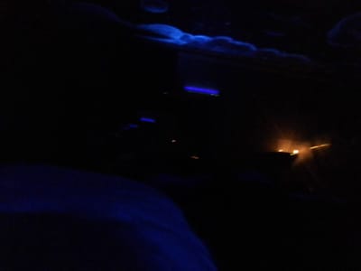 The image depicts a low-light scene within a contained environment, likely a vehicle or interior space, at night in Long Beach, United States. The upper left quadrant features a diffused blue light illuminating a textured, undulating surface. In the mid-left, several distinct linear blue light sources are visible, consistent with electronic displays or indicator lights. The right side of the frame contains multiple prominent orange-yellow point light sources, suggesting external illumination from streetlights, vehicle lights, or distant structures viewed through a window. The lower foreground is occupied by a large, indistinct dark blue object or surface, possibly a seat or upholstery. No individuals, specific actions, or interactions are discernible.