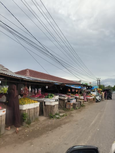 This image depicts a vibrant roadside fruit market in Grong-Grong, Indonesia, under a heavily overcast sky. The scene is viewed from a low angle, possibly from within a vehicle, with a car's dashboard or mirror visible in the bottom right.

Along the left side of a paved road, numerous market stalls are set up. These stalls feature simple structures with corrugated metal and red-tiled roofs, providing shelter for vendors and their goods. The stalls are abundantly stocked with a variety of fresh fruits, including bright orange tangerines or oranges, red apples, large watermelons, vibrant pink dragon fruit, and bunches of red grapes, all displayed in white plastic crates and rustic wooden boxes.

Several people are present in the market. In the immediate foreground on the left, a person dressed in a long, dark brown robe or dress is partially visible, their back to the viewer, standing near a fruit display. Further down the road, a group of people, including some women wearing headscarves, are gathered near a dark pickup truck parked on the side of the road. A white scooter is also parked nearby. The overall activity suggests a busy market day, with people shopping or tending to their stalls.

Above the market, a dense network of black electrical or utility wires stretches across the grey, cloudy sky. In the mid-ground, a blue sign with partially obscured white text is visible, which appears to read "Z-R...Ribu Suci." The road itself is made of asphalt and appears to be in moderate condition. The image captures a slice of everyday life and commerce in a local Indonesian setting, characterized by the informal market structure and overhead power lines.