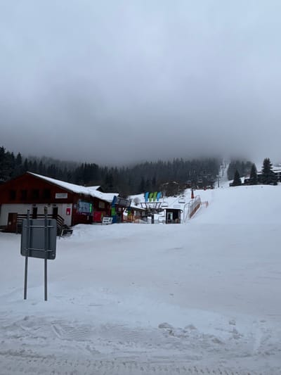 A snowy winter scene features a ski resort building with a red roof, surrounded by trees and a ski lift. The visibility is reduced due to heavy fog and snow. The foreground showcases a ski slope covered in snow, with tracks from previous skiers. The landscape is dominated by white and gray tones.