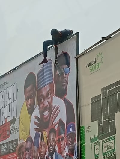 In Kofar Mazugal, Nigeria, an outdoor scene captures a dark-skinned male positioned precariously atop a large billboard against an overcast gray sky. He is clad in a dark, long-sleeved shirt with subtle orange accents and dark trousers, appearing barefoot on his right foot. The individual lies horizontally, stomach-down, across the top metal frame of the billboard. His right leg is bent, with his foot resting on the frame, while his left leg extends backward. His left arm reaches downward onto the billboard's surface, his hand gripping an unseen object or area, suggesting an action of repair, installation, or tampering. His right arm is bent with the hand near his head.
The billboard beneath him advertises an entertainment event, displaying "AWARD BAND ENTERTAINMENT," partially visible text "R!N RIYA," "MULTIPLE LAFFS," and "2nd + Coming," along with "HILTON PALACE HOTEL." It features multiple illustrated male figures, with a central smiling figure wearing a white garment and a striped cap, hands clasped.
To the right, a second billboard is visible, advertising "vertical Solar." Its face displays the "vertical Solar" logo in green text with a sunburst icon, alongside illustrations of solar panel arrays and "vertical Solar" branded tubular batteries. Both billboards feature dark, visible metal framing.