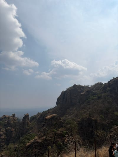 The image captures a scene in Tepoztlán, Mexico. A large, rocky mountain rises on the right, covered with sparse vegetation. A fence runs along the edge of the mountain. The sky takes up the left side of the frame, filled with white and gray clouds. A few people are visible in the upper right corner of the image.