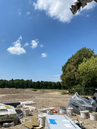 This image depicts a construction site under a clear blue sky with scattered white clouds. In the foreground, various construction materials are visible, including bags of what appears to be cement or plaster, white foam insulation pieces, and several buckets, some of which contain remnants of paint or plaster. A large, clear plastic-wrapped bundle of materials is also present. The ground is covered in gravel and dirt, with piles of excavated earth or rubble in the mid-ground. A line of lush green trees forms a backdrop, suggesting an outdoor, possibly rural or semi-rural, setting. In the upper right corner, a person's foot wearing a shoe is visible, seemingly resting on something above the frame, implying the photographer is positioned at an elevated viewpoint. The scene is indicative of ongoing construction work. The specific location in Hückelhoven, Germany, is not discernible from the visual elements alone, but the presence of construction suggests a developing area.