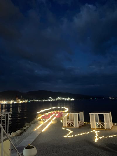 The image depicts a nighttime scene at a seaside location, likely a resort or beach area, in Sementsovka, Russia. The sky is dark and cloudy, with a faint glow on the horizon suggesting distant city lights and possibly port facilities. In the foreground, a pathway is illuminated by strings of warm white fairy lights that wind along the ground and around two white, open-sided cabanas. The cabanas appear to be furnished with seating. Further along the path, more fairy lights outline rows of what seem to be seating arrangements, possibly chairs or benches, with some appearing to be a vibrant pink color. The ground is covered with small stones or gravel. The sea is calm and reflects the lights from the distant shoreline, which is characterized by a dark silhouette of hills or mountains. The overall atmosphere is serene and inviting, with the lighting creating a festive and romantic ambiance.