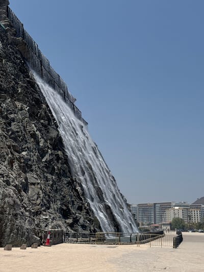 This image displays the artificial waterfall at the Khor Fakkan Corniche in Khor Fakkan, United Arab Emirates, under a clear, bright blue sky. The prominent feature is a large volume of water cascading down a steep, dark grey, rugged rock face that dominates the left half of the frame, flowing from top to bottom. The waterfall appears multi-tiered, with water breaking into numerous streams as it descends, creating a foamy appearance at its base.
In the foreground, a wide, light-colored paved area, possibly a pedestrian promenade, extends across the lower portion of the image. A black and yellow metal fence with curved sections borders this paved area, separating it from the waterfall's collection basin and a roadway beyond. Near the base of the rock face, on the left, are three rectangular concrete blocks, a stone structure with a small entrance, and a red and white traffic cone. White Arabic text, resembling "تسلل", is inscribed on the rock face above these objects.
In the background on the right side of the image, several multi-story modern buildings with light-colored facades and numerous windows are visible, indicating an urban or resort area. A few vehicles are discernible on a road behind the fence and in front of the buildings, and a small number of indistinct human figures are present on the paved area and near the road. A distant mountain range is visible on the far right horizon.