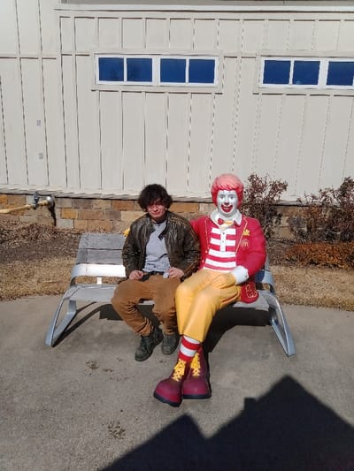 A person with dark hair and a brown jacket sits on a gray bench next to a Ronald McDonald statue. The statue is wearing a red suit, yellow pants, and large red boots. The background includes a building with small rectangular windows and a small area of dry bushes. The sun casts a shadow near the bottom of the frame, adding depth to the scene.