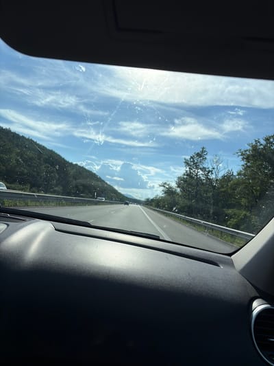 The image is a view from the interior of a car, looking forward through the windshield. The car is traveling on a multi-lane highway. On the left side of the highway, there is a steep, tree-covered hill with a metal guardrail separating the road from the hillside. On the right side of the highway, there are more trees. The sky is blue with white, wispy clouds and a bright sun is visible, causing some glare and reflections on the windshield, which also has visible smudges and streaks. Several cars are visible on the highway, traveling in the same direction as the car from which the photo is taken. The dashboard of the car is visible in the foreground. The scene appears to be in a rural or mountainous area. Based on the provided context, the location is the city of Novi han, Bulgaria.