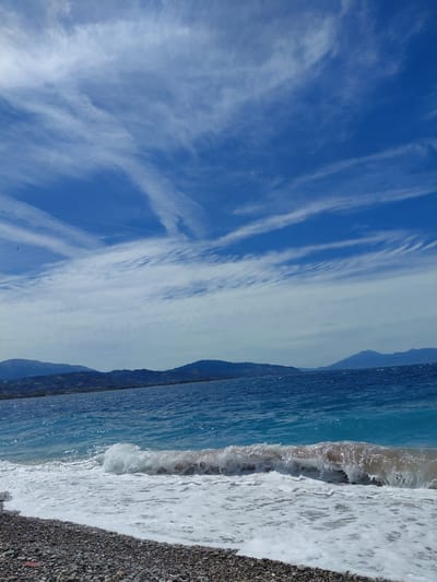 A pebble beach meets the turquoise waters of the sea under a bright blue sky dotted with wispy white clouds. In the distance, hazy mountains form a distant horizon. The foreground is dominated by the pebble-strewn shore and the foamy crest of a wave breaking onto it. A small, dark bird is visible in flight in the upper left portion of the sky. The scene suggests a tranquil day at the seaside in the Municipal Unit of Aegio, Greece.