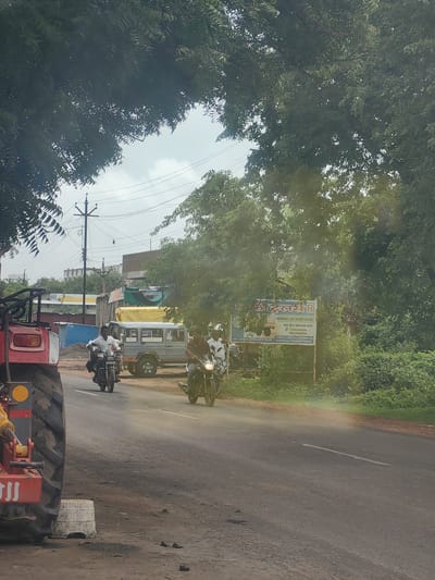In Malkapur, India, a busy road is captured under a canopy of lush green trees. The scene unfolds with a red tractor in the foreground, its large tire and metallic components prominent on the left side of the frame. A dusty, grey asphalt road stretches into the distance, marked with a white dividing line.
On the road, several individuals are actively engaged in travel. Two men are riding motorcycles, one in front of the other. The lead motorcyclist, wearing a white shirt, is seen from the side, while the second motorcyclist is partially obscured. To their right, a light grey utility vehicle with tinted windows is parked or moving slowly.
Further down the road, a white and yellow sign stands near a dense patch of green foliage. The sign displays text in Hindi, with some numbers visible, suggesting it is an advertisement or informational board. A utility pole with multiple wires is also visible in the background, indicating the presence of infrastructure. The overall environment suggests a rural or semi-urban setting in India, with a mix of vehicles, vegetation, and signage.