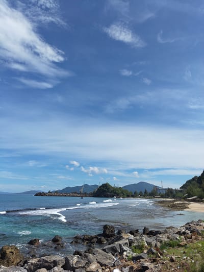 The image depicts a coastal scene in Naga Umbang, Indonesia. In the foreground, large, dark rocks are scattered along the shoreline with some sparse vegetation growing amongst them. The turquoise-colored water has gentle waves lapping at the rocky beach. Further out, the water becomes a deeper blue.

Across the bay, a breakwater constructed of dark rocks stretches into the water. Beyond the breakwater, the coastline features lush, green hills and mountains. Two cranes stand on the shore, hinting at industrial or port activity. A building and tower are visible among the trees on the coast. The sky is a bright blue with scattered white clouds. The scene is well-lit with sunlight.