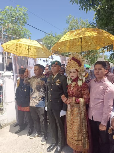 A wedding procession is underway in Geulangganggampong, Indonesia, under a bright, sunny sky. The bride, adorned in a traditional red and gold outfit and an elaborate headdress, is accompanied by the groom, who is in military uniform. They are walking forward, holding hands. Two large, ornate yellow umbrellas are held above them, providing shade. A crowd of onlookers, including men in traditional attire and women in headscarves, follows behind. In the background, a banner with text, including the word "WEDDING," is visible. The setting appears to be outdoors, with a white wall and some vegetation visible.