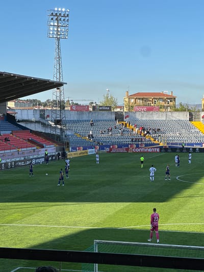 The image shows a soccer match at Estádio Municipal Engenheiro Manuel Branco Teixeira, located in Chaves, Portugal. Players in white and navy uniforms are on the green field, along with a goalkeeper in a pink jersey with the number 23. On the sidelines, branding and advertisement banners are visible. The stadium seating consists of blue and red sections, with a few people in the stands. Behind the stadium, buildings and trees are present. The weather is sunny, and a large stadium light fixture stands tall on the left.