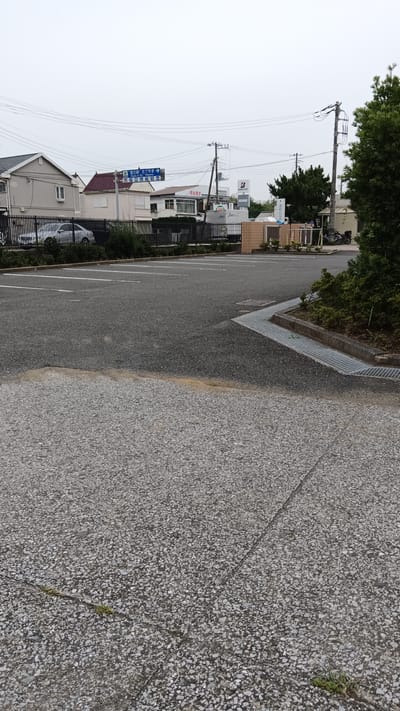This is a ground-level, full shot of a paved parking lot in Tateyama, Japan. In the background, a blue road sign indicates directions to "Tateyama Station" and "Tateyama-ko" (Tateyama Port). Businesses like "Bridgestone" are visible, as are residential buildings and a utility pole. The parking lot is mostly empty, with a few cars parked along the left side. A metal grate runs along the edge of the parking lot, indicating a drainage system. The sky is overcast, and the overall scene is muted in color.