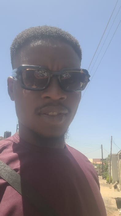 A male individual is depicted in a close-up, outdoors, under a clear, bright blue sky in Badiko, Nigeria. The subject, an adult male with short, dark, tightly curled hair, occupies the majority of the frame, facing forward. He wears dark, rectangular sunglasses with a mottled frame, and a plain, short-sleeved, maroon crew-neck shirt. A dark strap is visible diagonally across his left shoulder. His mouth is slightly open, revealing his upper front teeth.
In the background, to the subject's left, a dark water storage tank is visible on a distant rooftop. To the subject's right, a narrow, unpaved street or alley recedes into the distance, lined with low-rise buildings and some green vegetation. Utility poles with multiple overhead wires are present along the street. The environment appears sunlit, indicating daytime conditions.