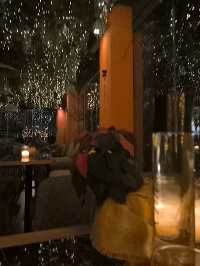 The image depicts a dim, ambient scene at an evening establishment in Palaio Faliro, Greece, likely a restaurant or lounge. The time of day is night, with the primary illumination coming from countless small, warm-toned string lights suspended above, possibly on trees or foliage, creating a sparkling, festive canopy.

In the immediate foreground, a dark, reflective tabletop, possibly marble, is visible. On the right, a tall, clear glass or bottle stands with reflections of the ambient lights. To its left, a textured yellow/brown object, possibly a woven bag or cushion, is partially obscured. Resting on what appears to be a chair or another surface are folded personal belongings, including dark clothes with a prominent red or pink section, and a brown item.

The midground features a prominent, tall, ochre or orange-colored pillar or wall section. Further into the scene, another table is visible with a lit candle, contributing to the intimate atmosphere. Glasses and perhaps other dinnerware are also on this table. Seating areas with upholstered chairs are faintly discernible around the tables.

The background reveals more of the sparkling lights, suggesting a large outdoor or semi-enclosed area. Reflections in glass panels imply a partition or wall, through which distant, blurred city lights or other illuminated structures can be seen. No people are clearly visible, but the setup indicates a space prepared for patrons.

The overall activity suggested is a relaxed evening out, such as dining or enjoying drinks. The ambiance is cozy and magical, attributed to the abundance of decorative lights. There is no visible text in the image.
