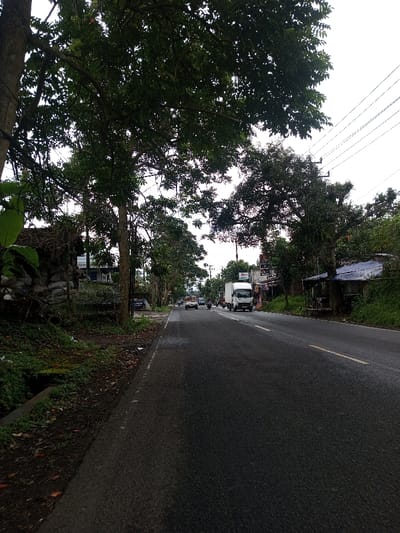 The image depicts a two-lane asphalt road in Kamji Na-Yekha, Bhutan, under an overcast sky. The road surface appears damp in sections, with a yellow solid line and a white dashed line delineating the lanes on the right side. On the left, a narrow paved shoulder borders a dirt and grass verge with a visible concrete drainage channel. Mature trees with dense green foliage line both sides of the road, their branches partially obscuring the sky and distant structures.
On the left side, behind the trees, a rudimentary structure constructed from stacked sacks (possibly sandbags) and tarpaulin is visible. Further down the road on the left, a blue building and other structures are present. Overhead, multiple power lines run parallel to the road.
On the right side, commercial or residential structures with some visible signage are interspersed among the trees and dense vegetation. A makeshift shelter with a blue tarp roof is also discernible.
Traffic is present on the road, moving away from the viewer. A white box truck occupies the right lane in the mid-ground. Behind it, several motorcycles or scooters, each with a rider, are observed between the lanes. Further in the distance, a light-colored car (possibly white or silver) is visible in the left lane, with other indistinct vehicles receding into the background. No pedestrians are clearly visible.